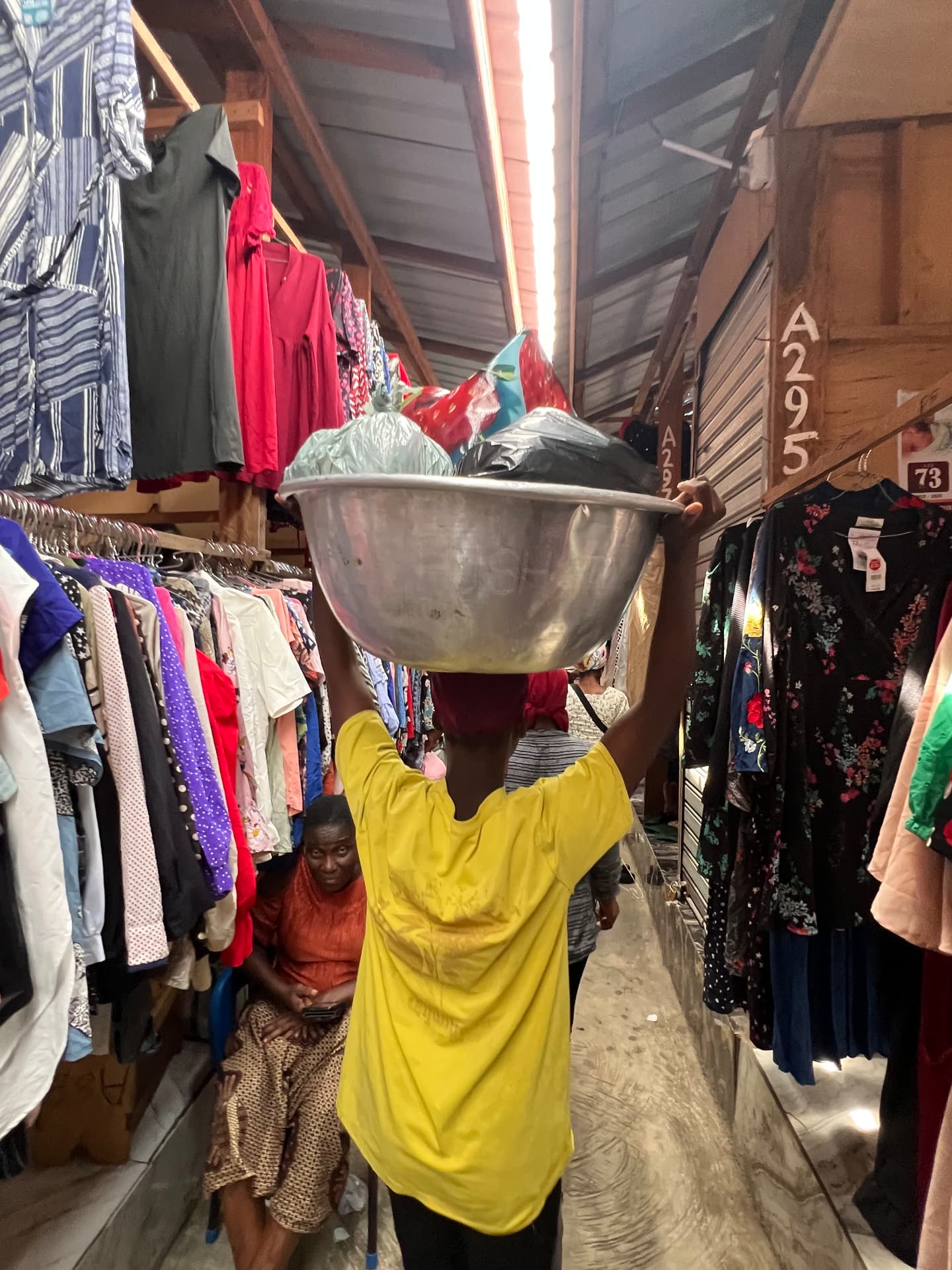 A kayayei porter carries a large aluminium bowl of goods on her head through a Kantamanto clothing aisle.