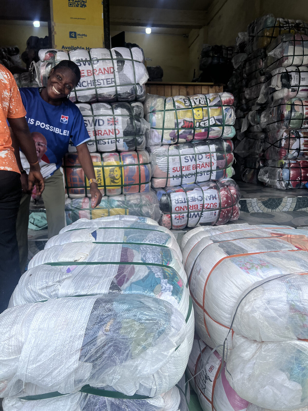 A trader smiling beside bales of imported second-hand clothing in a textile market